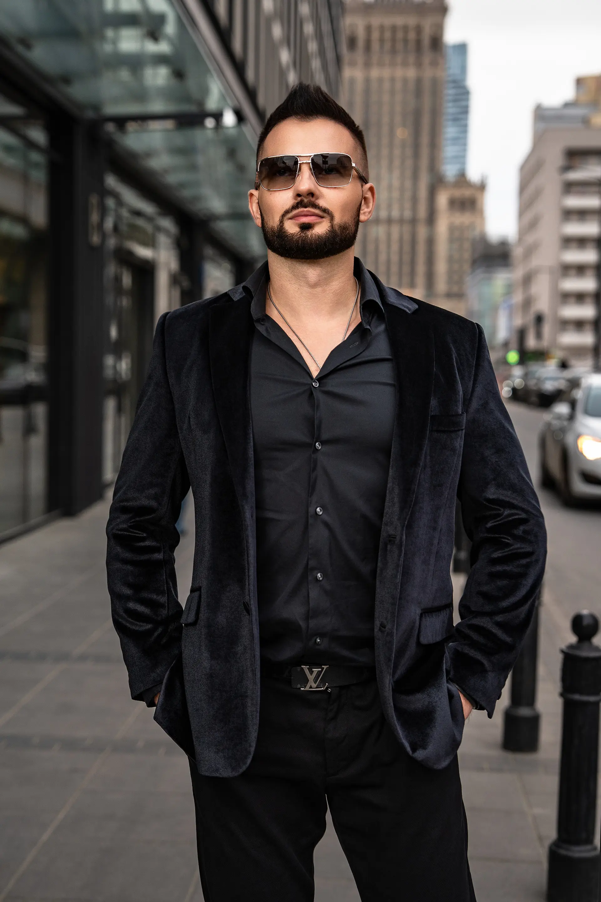 Grzegorz Nowak w czarnym garniturze i okularach przeciwsłonecznych na tle miejskiej zabudowy.