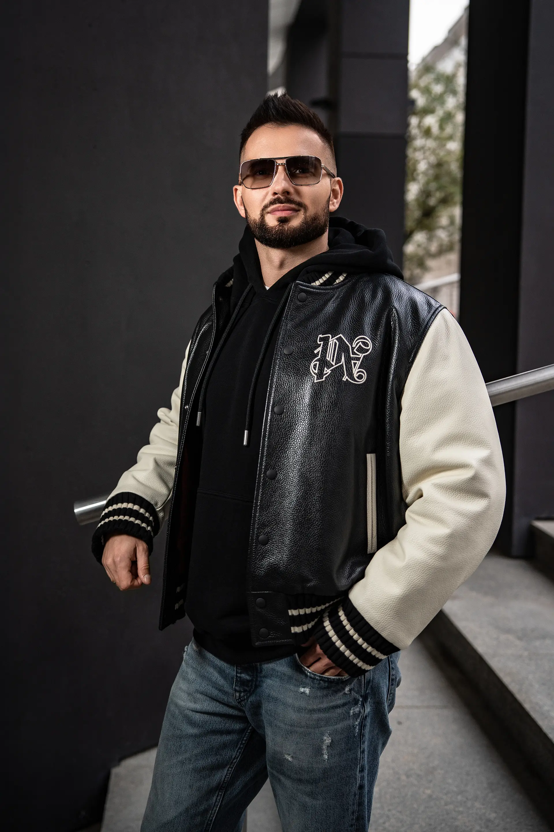 Grzegorz Nowak w kurtce varsity z białymi rękawami i okularach przeciwsłonecznych stojący przy nowoczesnym budynku.