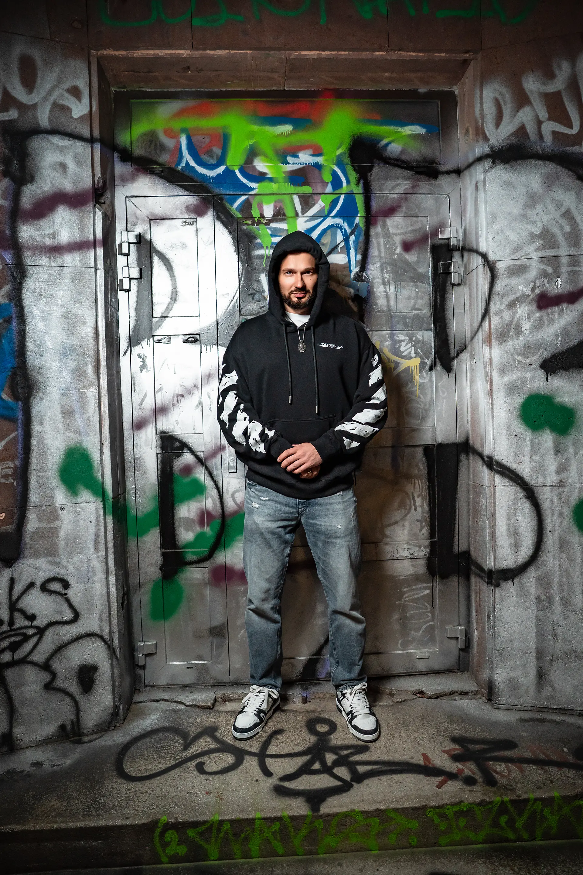 Grzegorz Nowak w czarnej bluzie z kapturem stojący na tle ściany pokrytej graffiti.