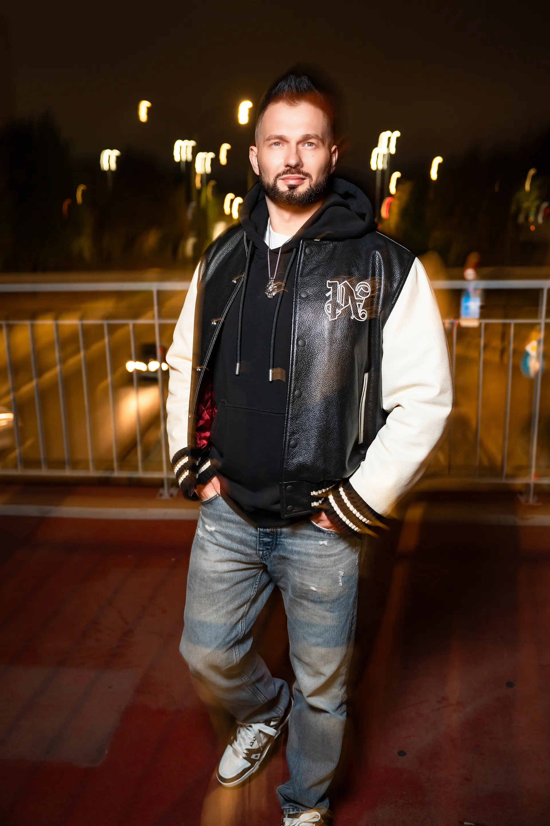 Grzegorz Nowak w skórzanej kurtce typu varsity i bluzie z kapturem pozujący na tle nocnego miasta.